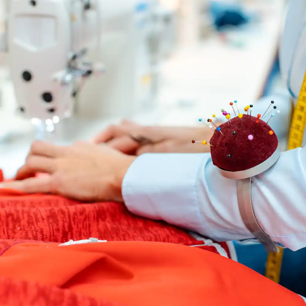 Ein Schneider näht roten Stoff an einer Nähmaschine, er hat ein Maßband umhängen und ein Nadelkissen am Arm